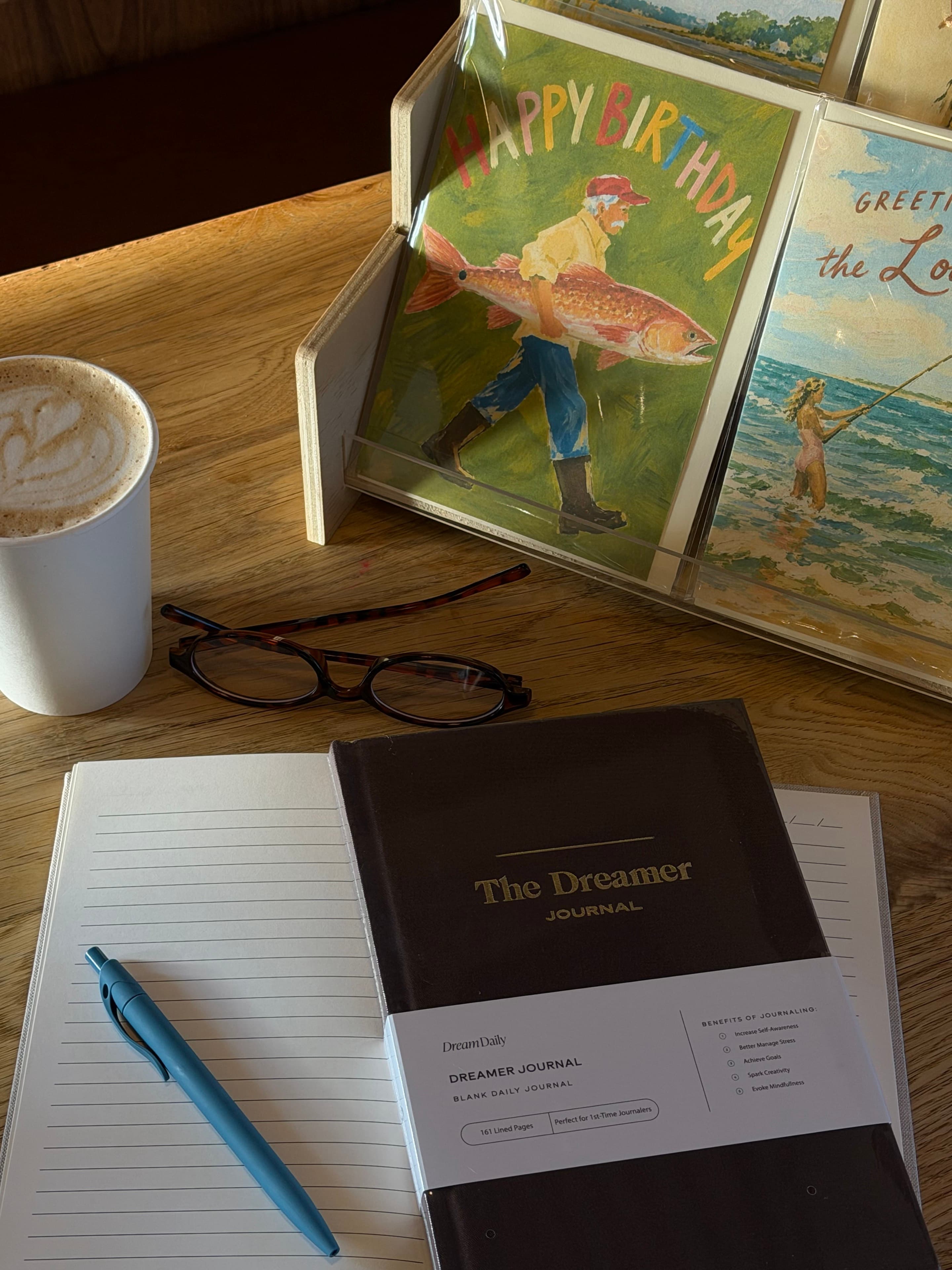 A Dream Daily journal, P.S. greeting cards, and a latte on a café table